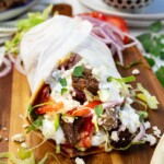 A Greek style pita bread stuffed with marinated lamb, feta, and salad and rolled up in baking paper, all on a wooden chopping board.