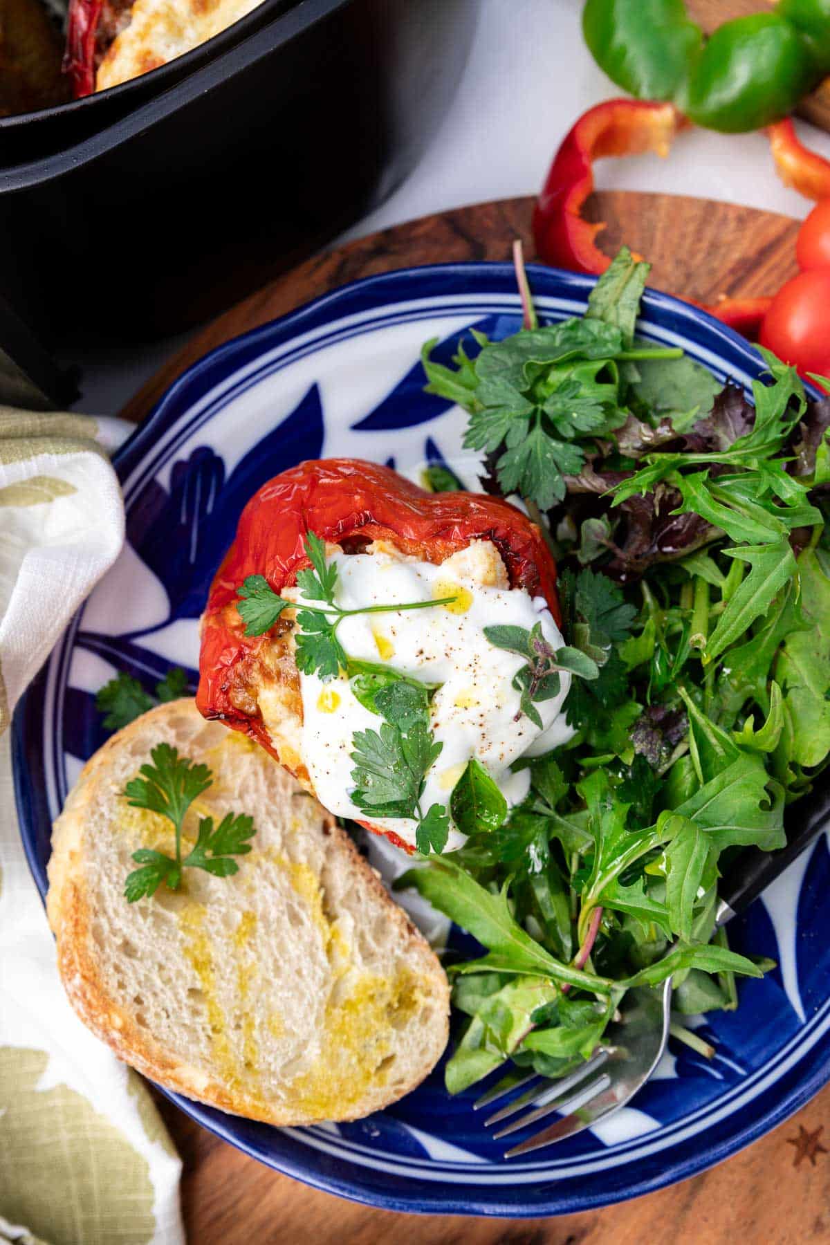 A patterned blue bowl with a stuffed red pepper with yogurt and herbs on top with green salad, a piece of bread and a fork, with ingredients, a green tea towel and an air fryer at the side.