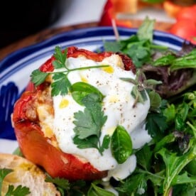 Closeup of a stuffed pepper with yogurt and herbs on top with green leafy salad, and a piece of bread at the side.