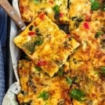 Overhead view of someone lifting a slice of baked vegetable frittata out of a lined sheet pan of it with a wooden handled knife and blue tea towel.