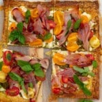 Overhead of a puff pastry tomato tart with prosciutto and basil on top, on baking paper and a wooden board with a knife.