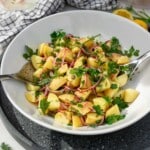 A big white dish of Greek potato salad on a grey ceramic tray with metal salad servers in it and with ingredients and a tea towel in the background.