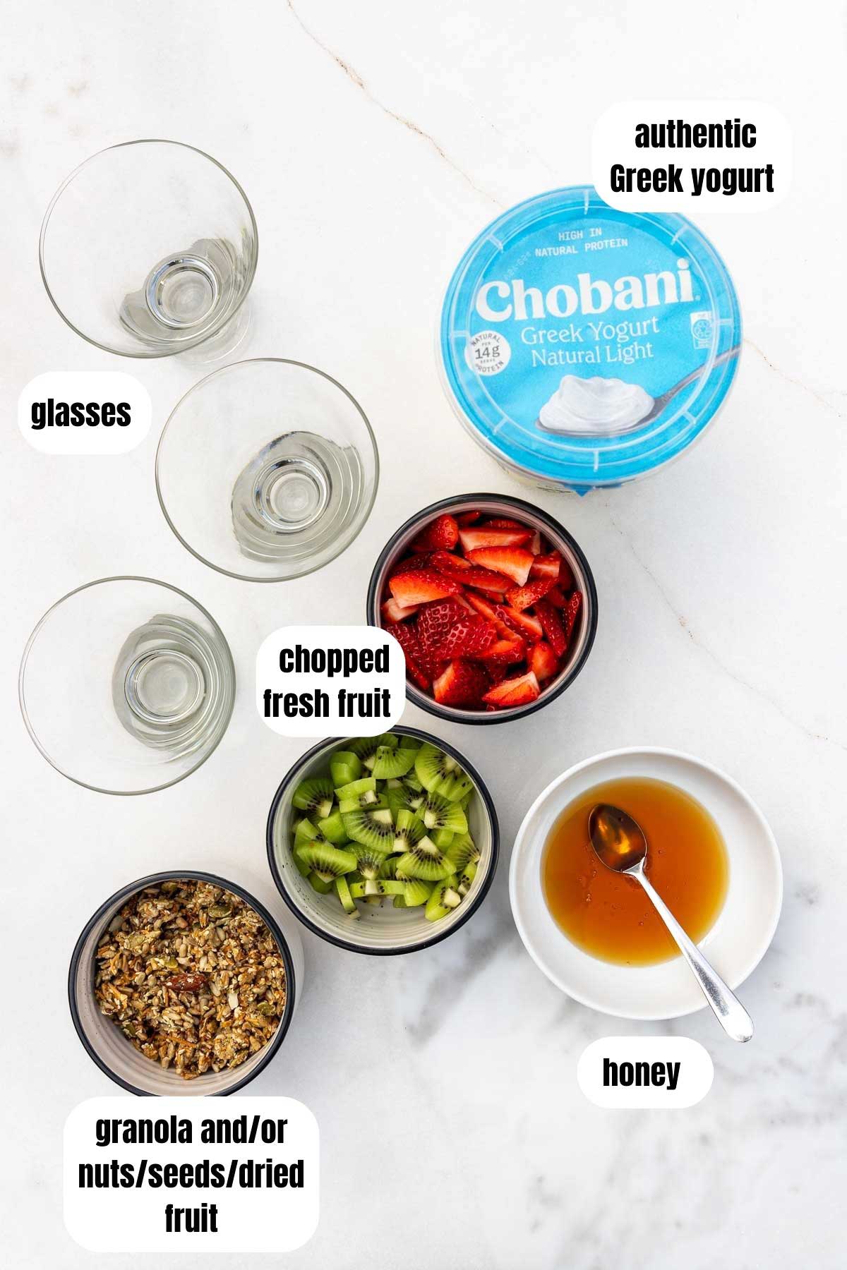 Overhead of all the ingredients to make Greek yogurt parfaits including clear glasses, authentic Greek yogurt, fresh fruit, honey, and granola.