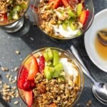 Overhead view of 3 Greek yogurt parfaits topped with honey, granola and chopped fresh strawberries and kiwi fruit on a grey ceramic tray and with spoons and ingredients at the side.