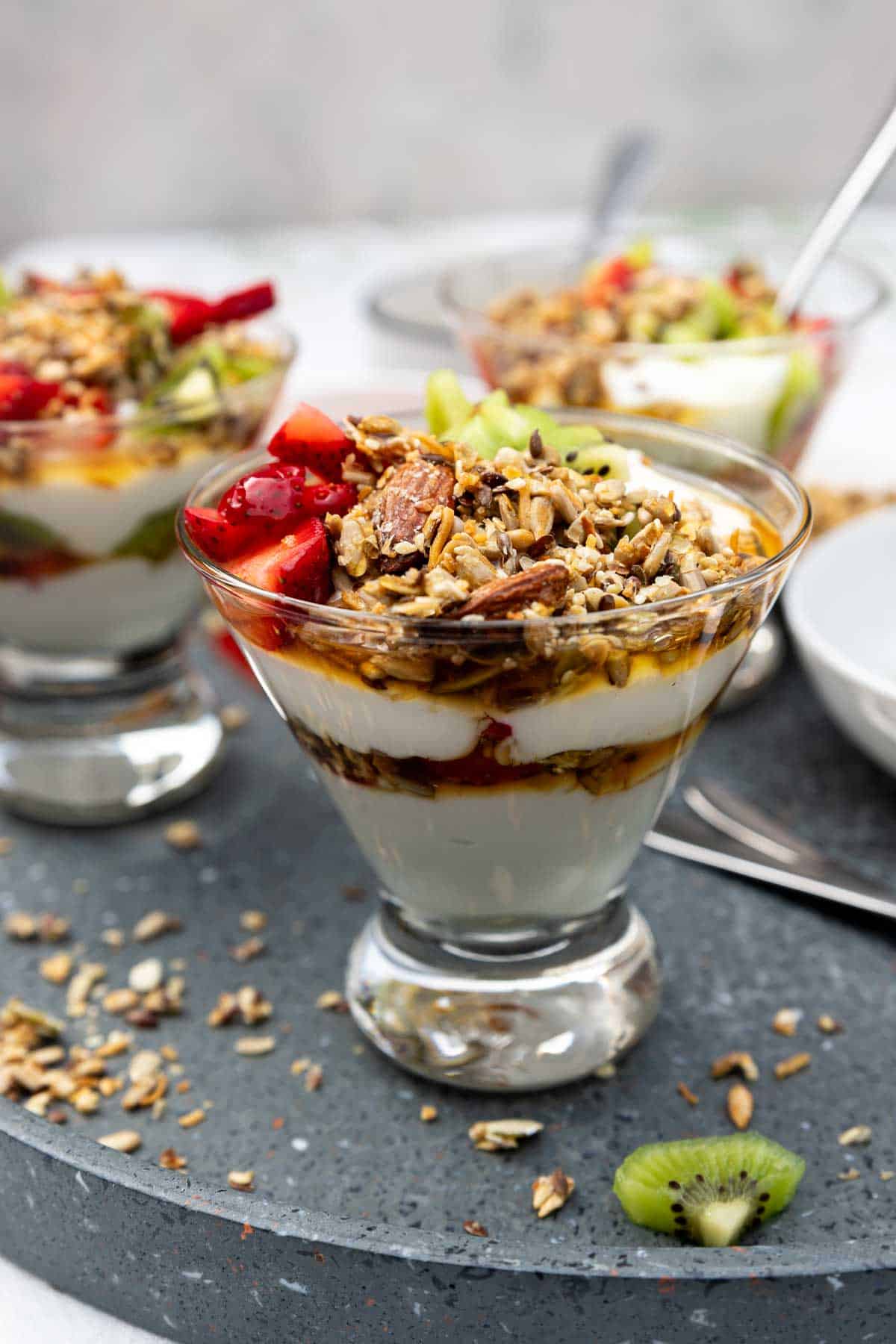 A dessert glass of Greek yogurt parfait with 2 layers of yogurt, honey, chopped fruit and granola, on a grey ceramic tray and with more parfaits in the background.