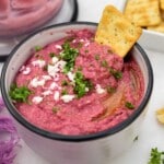 Closeup of the top of a bowl of beetroot and feta dip, with crumbled feta and fresh parsley on top, and with a cracker half dipped in.