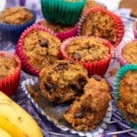 Carrot banana muffins in colorful muffin cases on a cooling rack on a purple tea towel with one ripped open to show the inside.