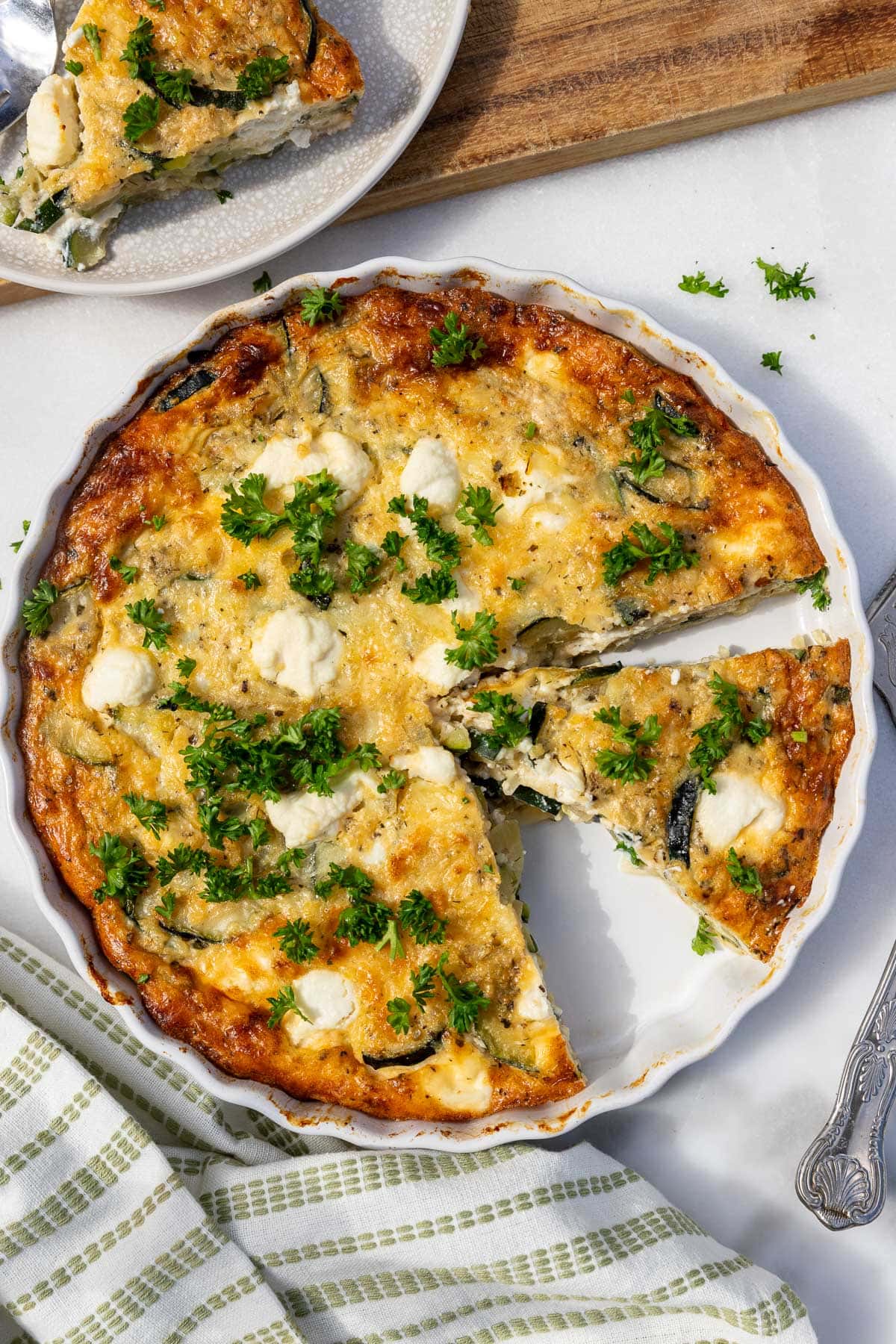 Overhead of a zucchini crustless quiche in a white quiche pan with a green and white tea towel and a small piece of quiche on a plate at the top.