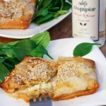 A close-up of a square feta filo parcel on a white plate with sesame seeds and honey sprinkled on top and a fork, with a bottle of ouzo in the background.