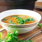 Thick red lentil soup in a white bowl with coriander on top and in front and with a spoon on a wooden board.