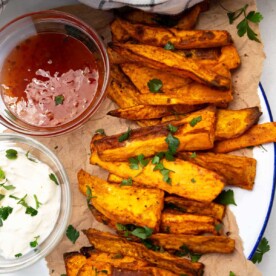 Overhead view of air fried sweet potato wedges on brown paper on a white plate with small bowls of sour cream and sweet chilli sauce to the left.