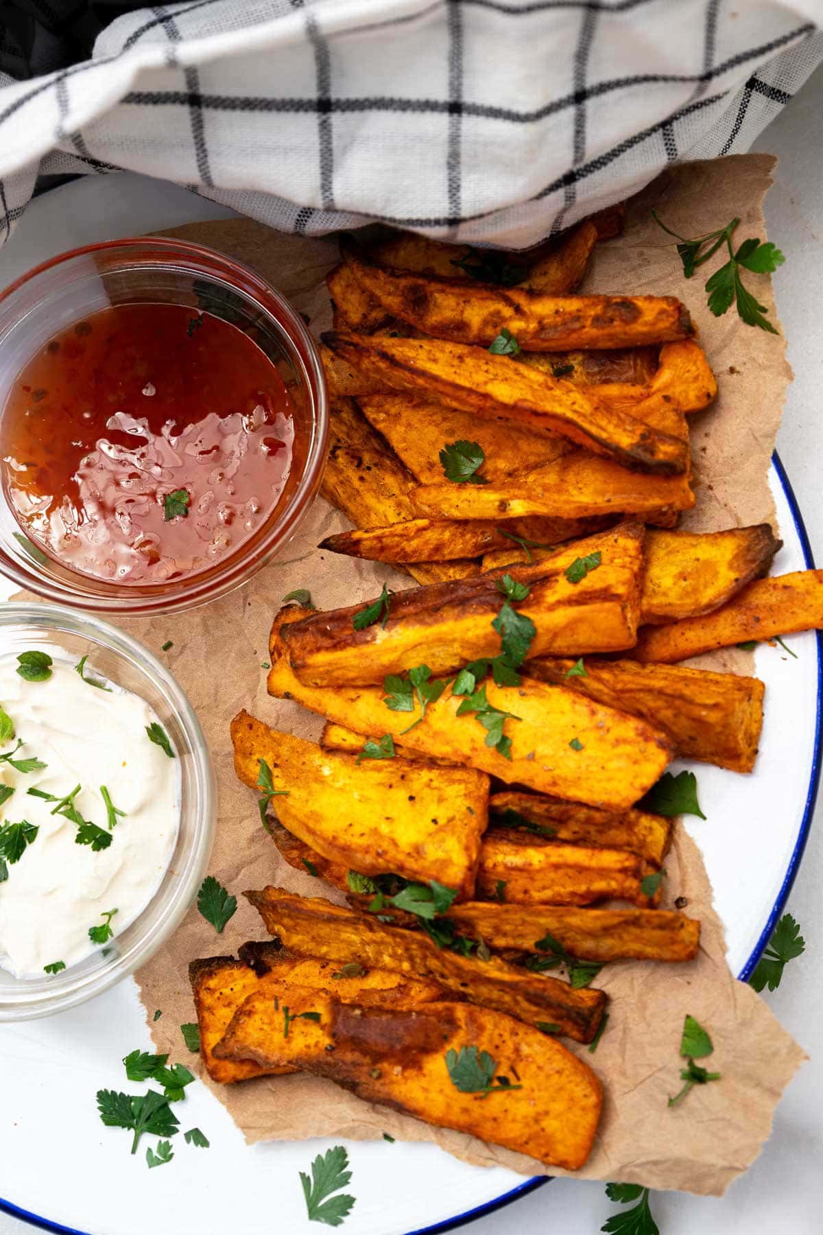 Overhead view of air fried sweet potato wedges on brown paper on a white plate with small bowls of sour cream and sweet chilli sauce to the left.