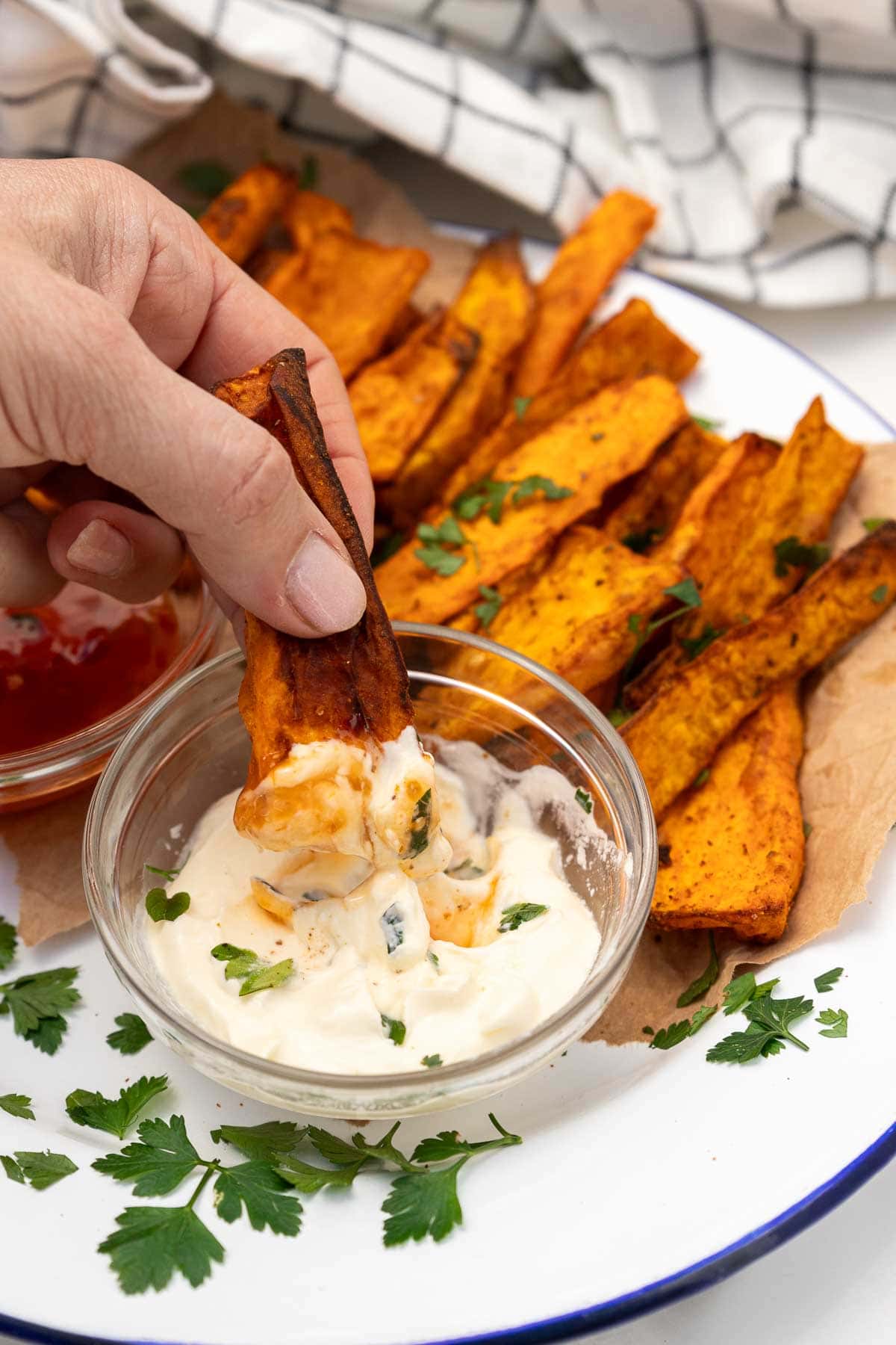 Someone's hand dipping an air fried sweet potato wedge into sour cream with sweet chilli sauce, on a blue rimmed white plate and with more sweet potato wedges in the background.