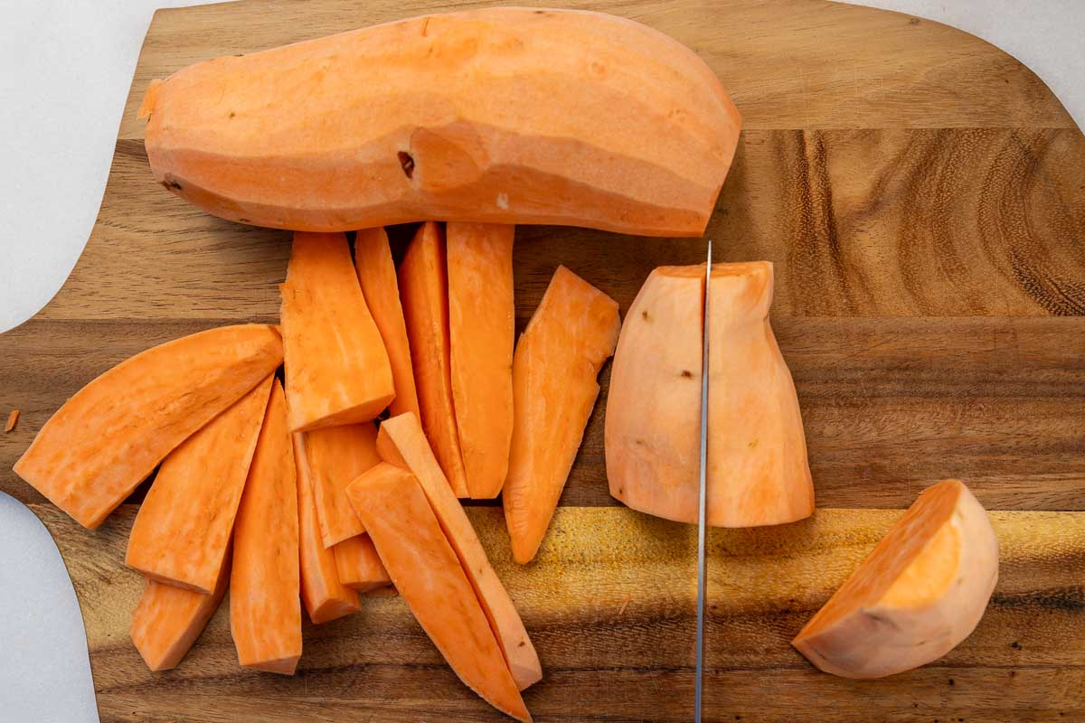 Overhead view of a knife slicing a piece of sweet potato in half, with raw sweet potato wedges at the side.