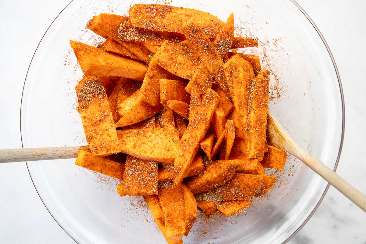 Overhead closeup view of a glass bowl of raw sweet potato wedges tossed with herbs and spices, with two wooden spoons in with them.