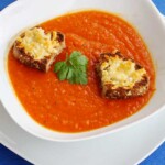 Closeup of a white dish of butternut squash and tomato soup with little cheese toasts on top on a bright blue background with a decorative spoon in front.