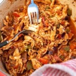 A closeup view of two forks shredding chicken breasts in sauce in a red dutch oven, with a red striped tea towel around the handle.