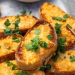 Closeup of a piece of grilled parmesan toast with fresh parsley on top and on top of a pile of more toasts, all on a lined grill pan.
