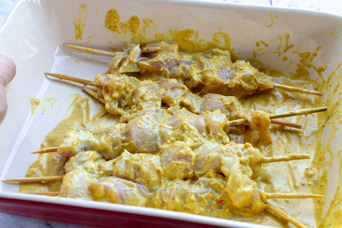 Closeup of raw chicken skewers smothered in a simple satay sauce in a red baking dish.