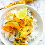 A white bowl of satay chicken with peanut sauce on a bed of rice and with twists of carrot and cucumber on top and a slice of lime.