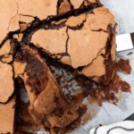 Extreme closeup of someone lifting up a big slice of Italian chocolate torta tenerina from the whole cake on baking paper and with a white and black striped tea towel.