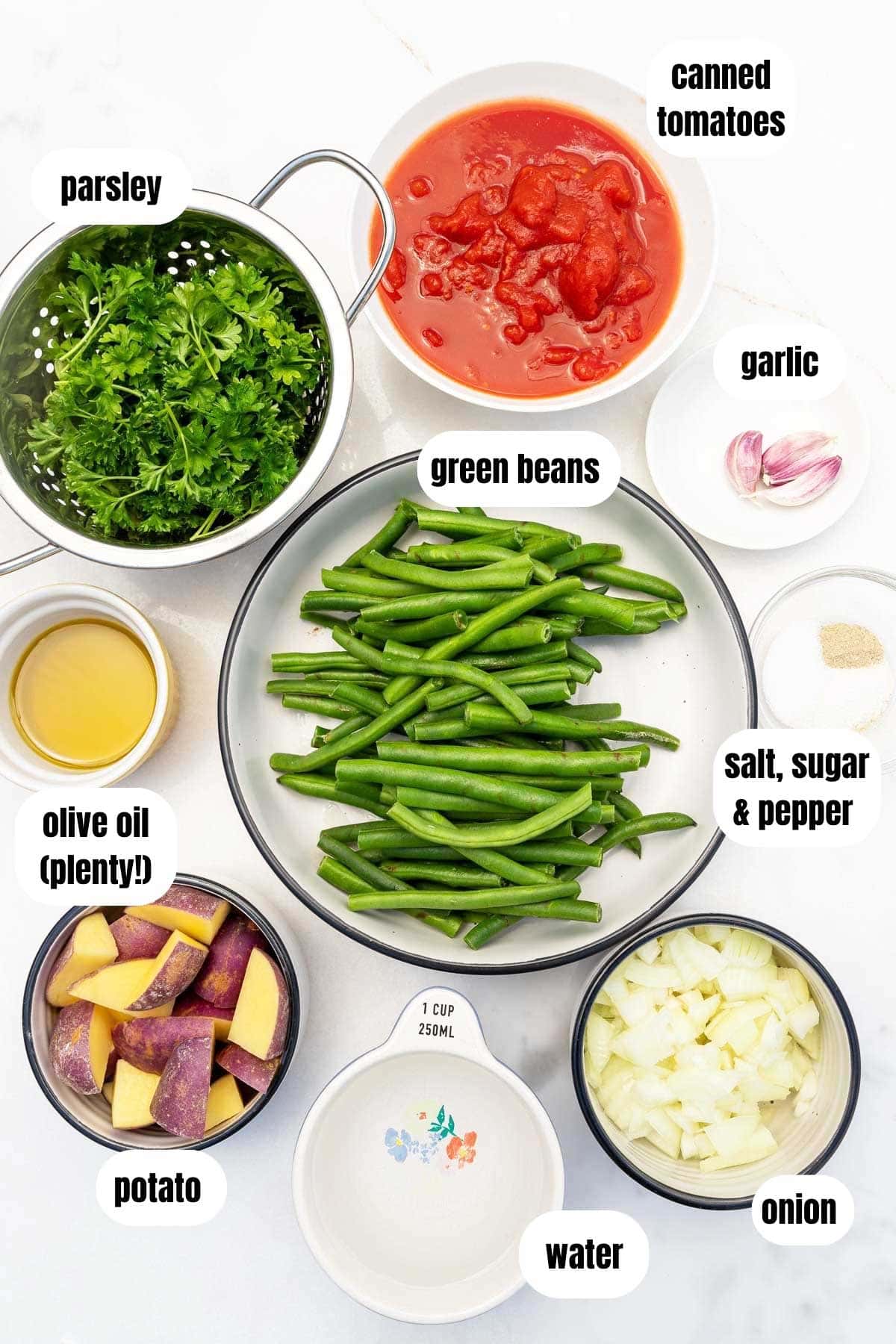 Overhead of all the ingredients needed to make fasolakia or Greek green beans including fresh green beans, chopped potatoes, chopped onion, garlic, lots of olive oil, parsley, canned tomatoes, water and salt, sugar and pepper.