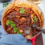 A Greek beef stew or stifado in a red cast iron pot on a wooden board with a blue tea towel and with bread and bowls in the background.