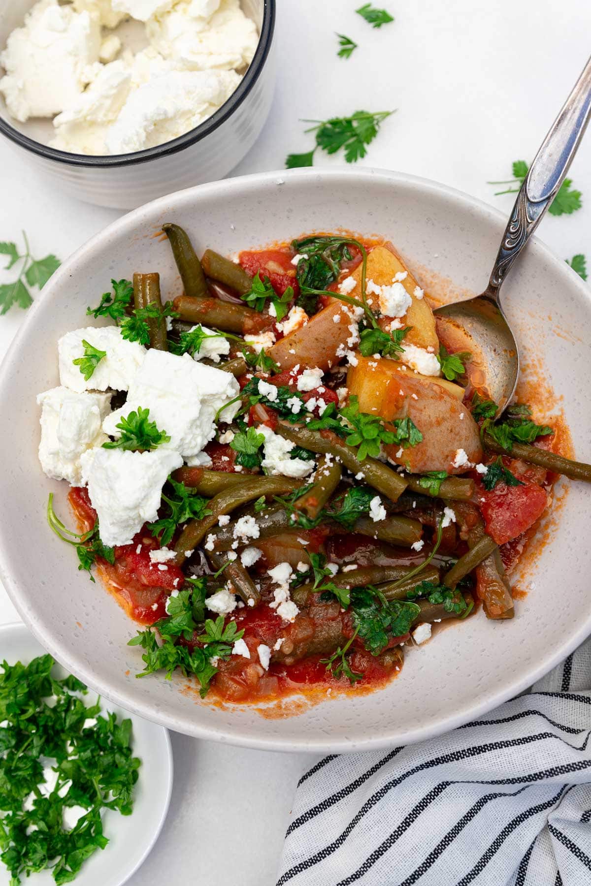Overhead view of a bowl of fasolakia or Greek green beans with a spoon in and feta cheese on top, with ingredients and a tea towel at the side.
