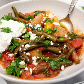 Closeup of a bowl of fasolakia or Greek green beans with a spoon in and feta cheese on top, with ingredients and a tea towel at the side.