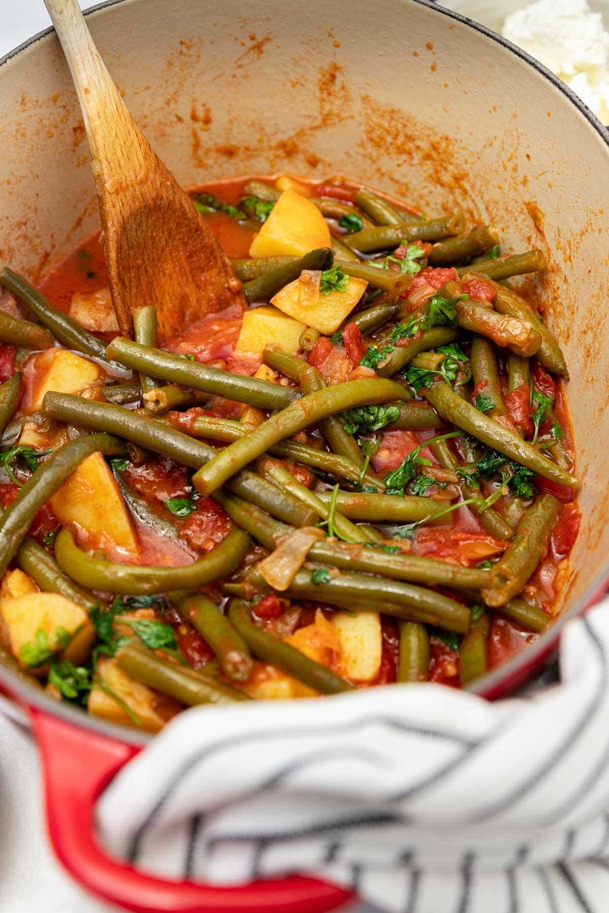 Closeup of a pan of cooked Greek green beans or fasolakia with a wooden spoon in it and a white and black tea towel at the front.