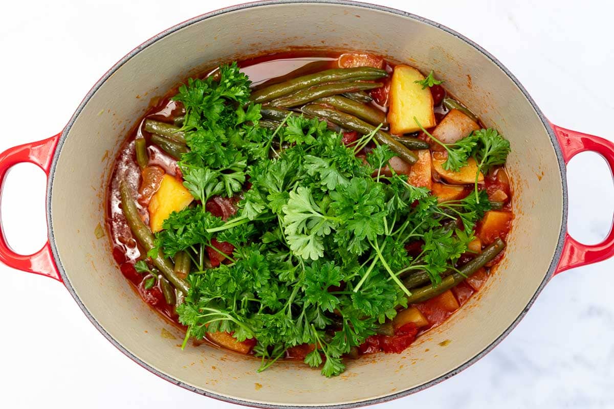 Overhead image of a red cast iron pot with a Greek green bean stew in it, with fresh parsley on top ready to be mixed in.