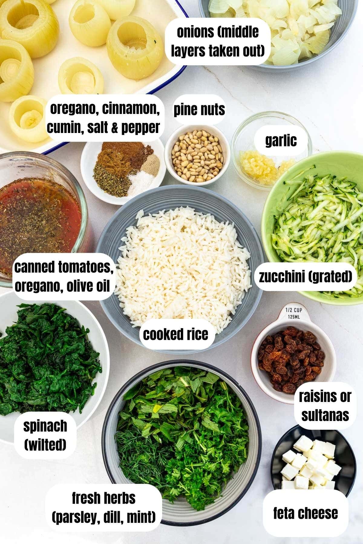 All ingredients needed to make stuffed onions including cooked rice, onions, fresh herbs, spinach, feta cheese, raisins or sultanas, grated zucchini, canned tomatoes, oregano, olive oil, spices, pine nuts and garlic.