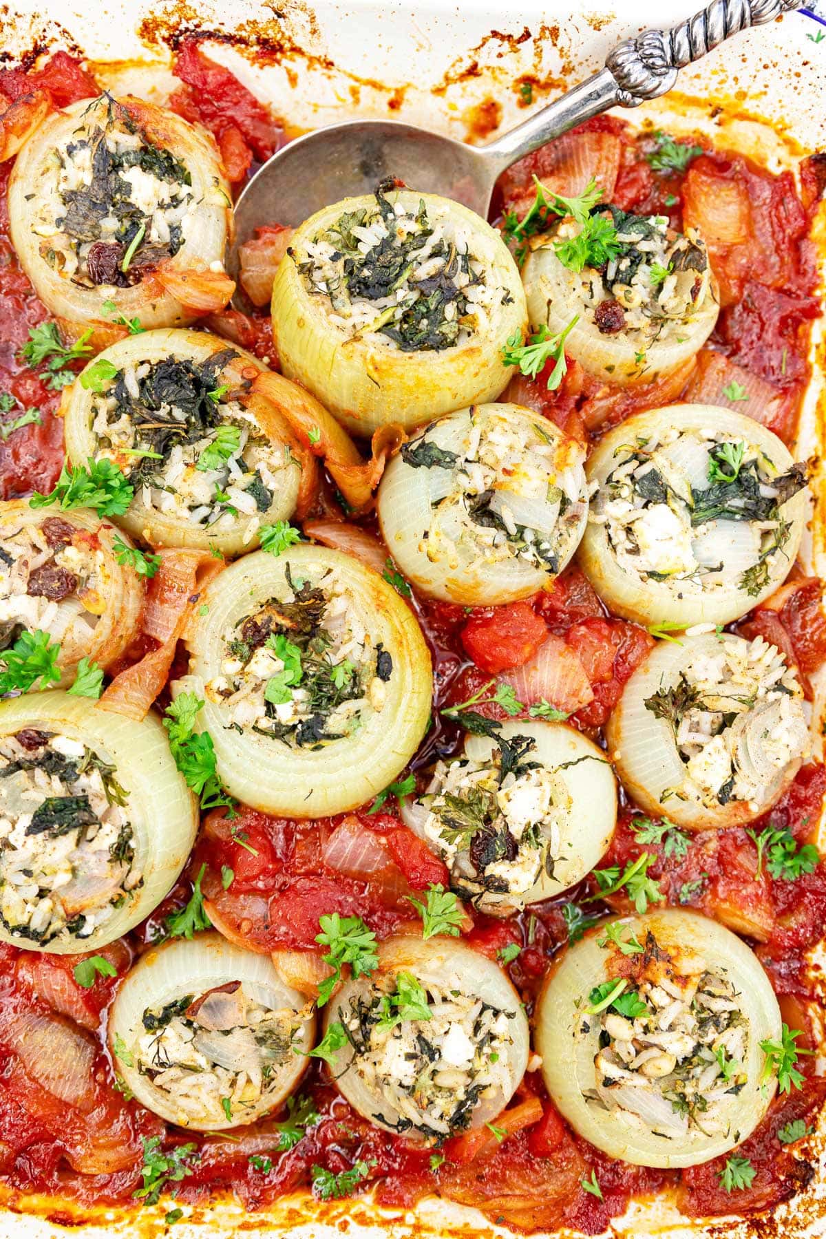 A pan of cooked Mediterranean style stuffed onions in a tomato sauce and with a decorative metal spoon in the pan.