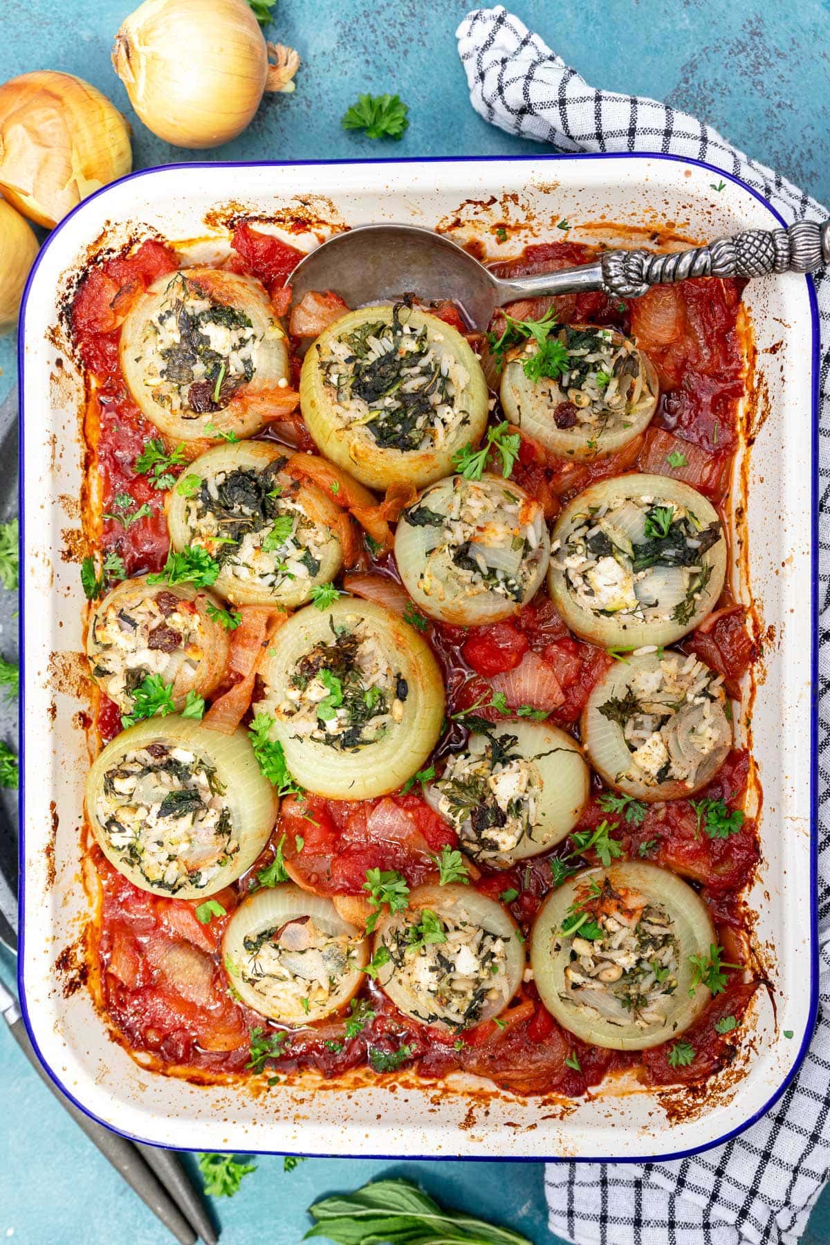Overhead view of a pan of cooked Greek style stuffed onions in tomato sauce on a blue background with checked tea towel and more onions in view at the top.
