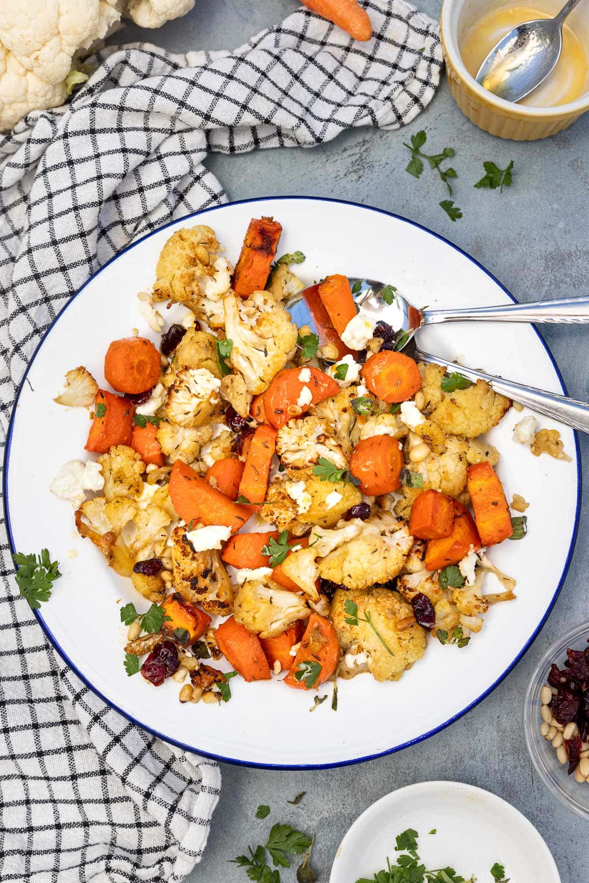 Roasted cauliflower and carrots on a blue rimmed white plate, scattered with feta cheese, cranberries, pine nuts and parsley, all on a blue background with checked tea towel.