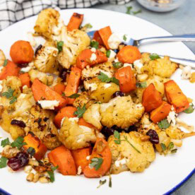 A blue-rimmed white plate with roasted carrots and cauliflower with cranberries, pine nuts, feta and parsley on top, on a pale blue background with a checked tea towel.