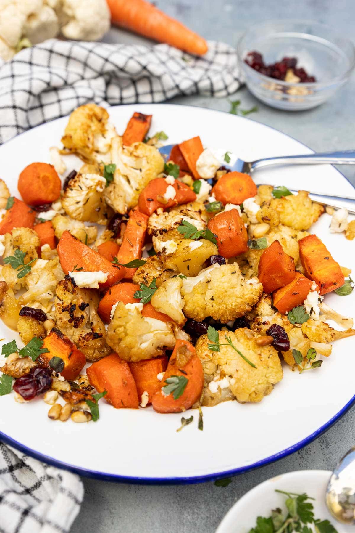 A blue-rimmed white plate with roasted carrots and cauliflower with cranberries, pine nuts, feta and parsley on top, on a pale blue background with a checked tea towel.