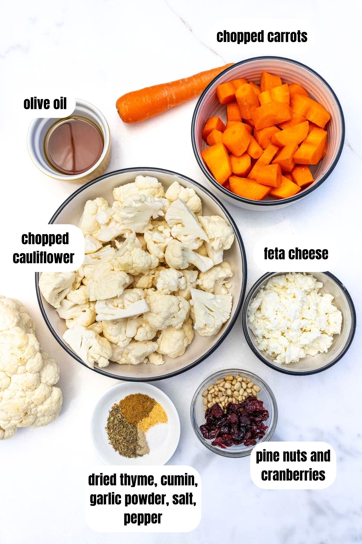 All the ingredients needed to make roasted cauliflower and carrots with cranberries and feta including cauliflower pieces, chopped carrot, dried thyme, cumin, garlic powder, salt and pepper, pine nuts and cranberries, feta cheese and olive oil.