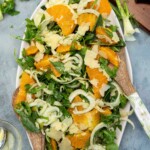 Overhead view of a fennel orange salad on a light beige platter with wooden salad servers on a pale blue background.