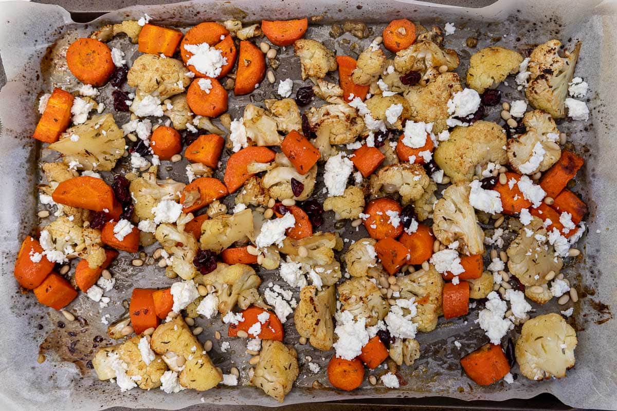 Roasted cauliflower and carrots on a lined baking sheet scattered with feta, pine nuts and cranberries.