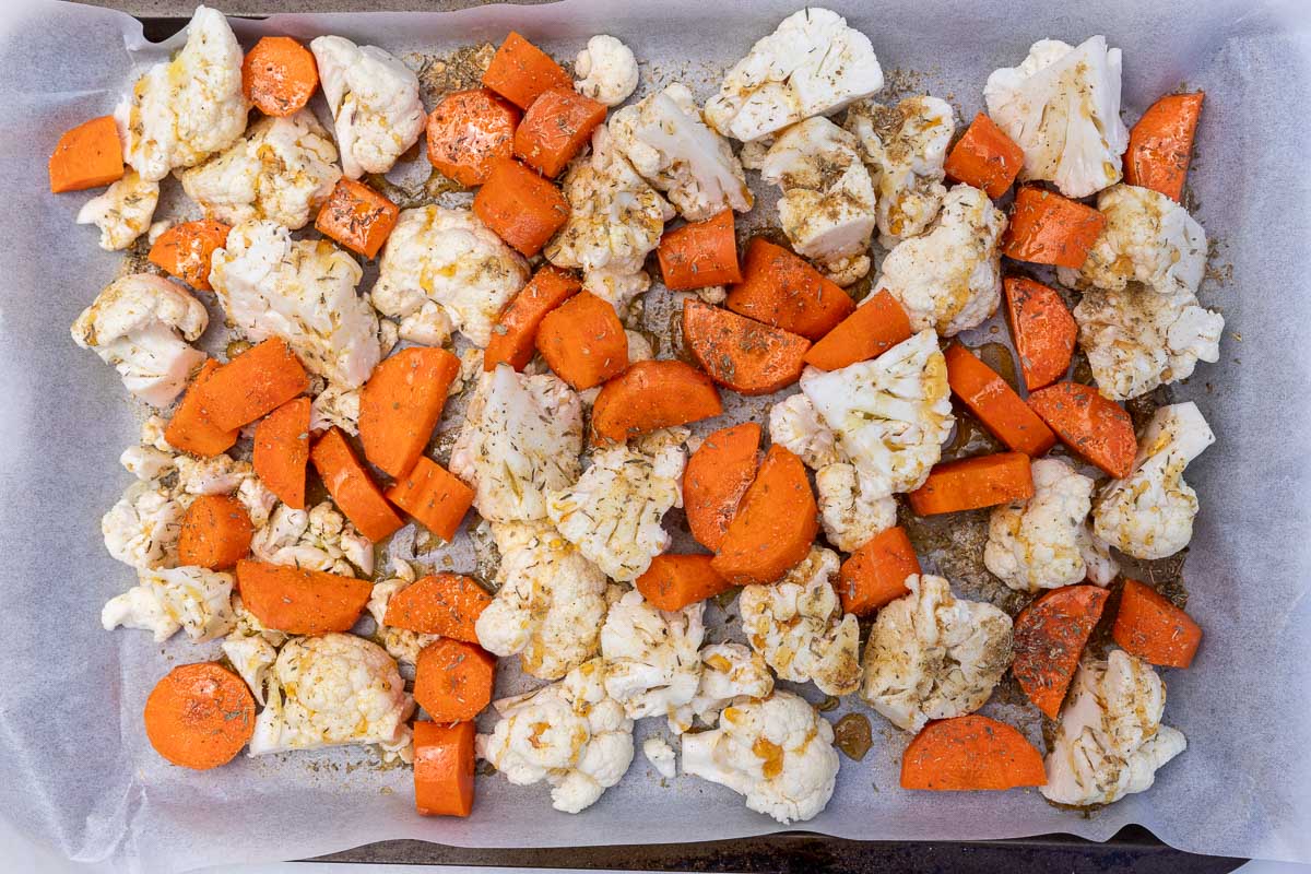 Pieces of raw cauliflower and carrots tossed with herbs and spices on a baking sheet lined with baking paper.