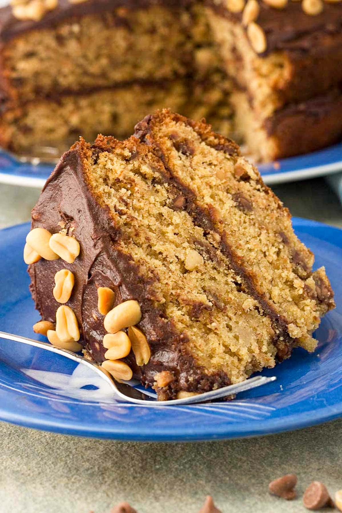 A closeup of a slice of peanut butter banana chocolate cake with chocolate icing and peanuts on top on a blue plate with a fork.