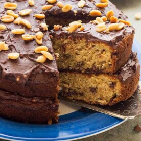 A closeup of a decorative cake slice taking a big slice of peanut butter banana chocolate cake with chocolate icing and peanuts on top out of a whole cake.