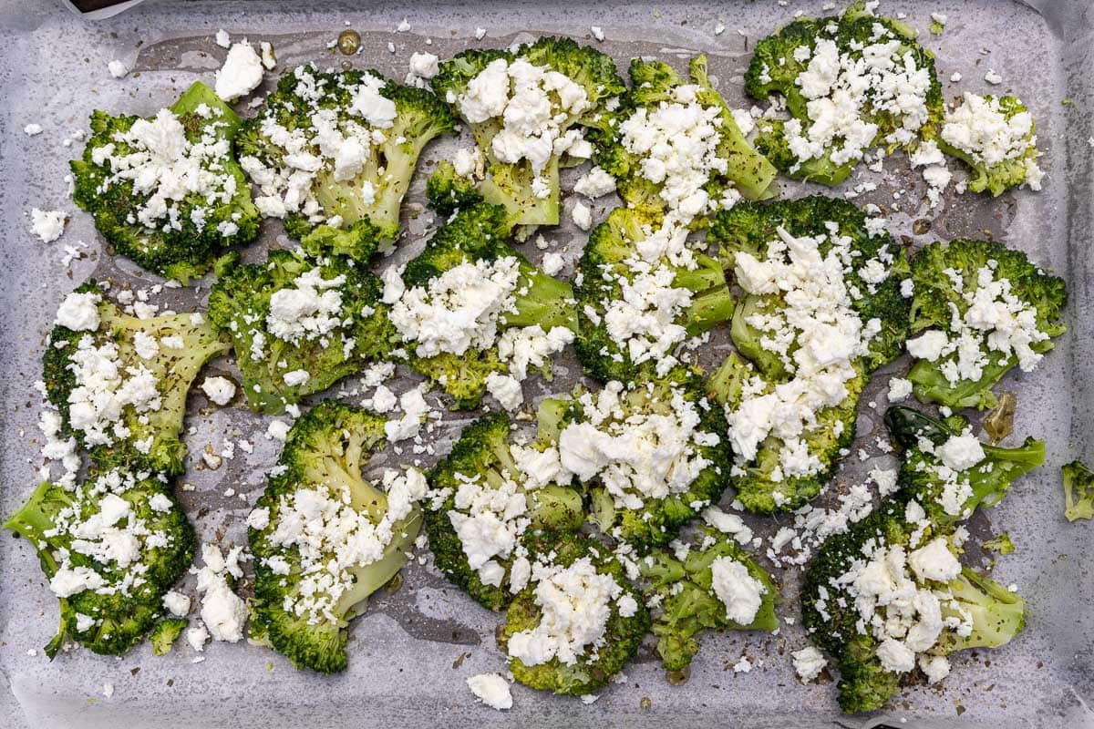Overhead view of parboiled smashed broccoli on a lined baking pan with lots of feta cheese sprinkled on top.
