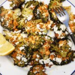Closeup view of a white plate with smashed broccoli and feta, a fork and a lemon segment on it, with the edge of a white and green tea towel.