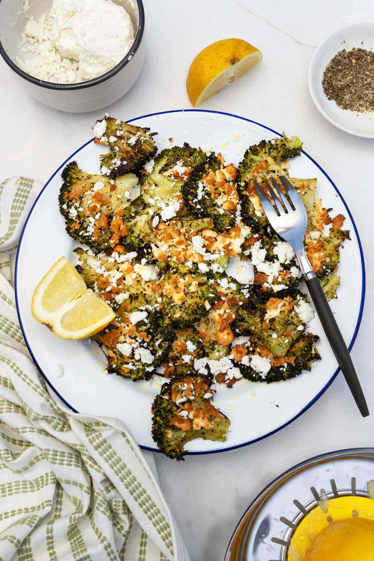 Overhead view of a white plate of smashed broccoli with feta with a fork, a green and white tea towel, and ingredients around it.