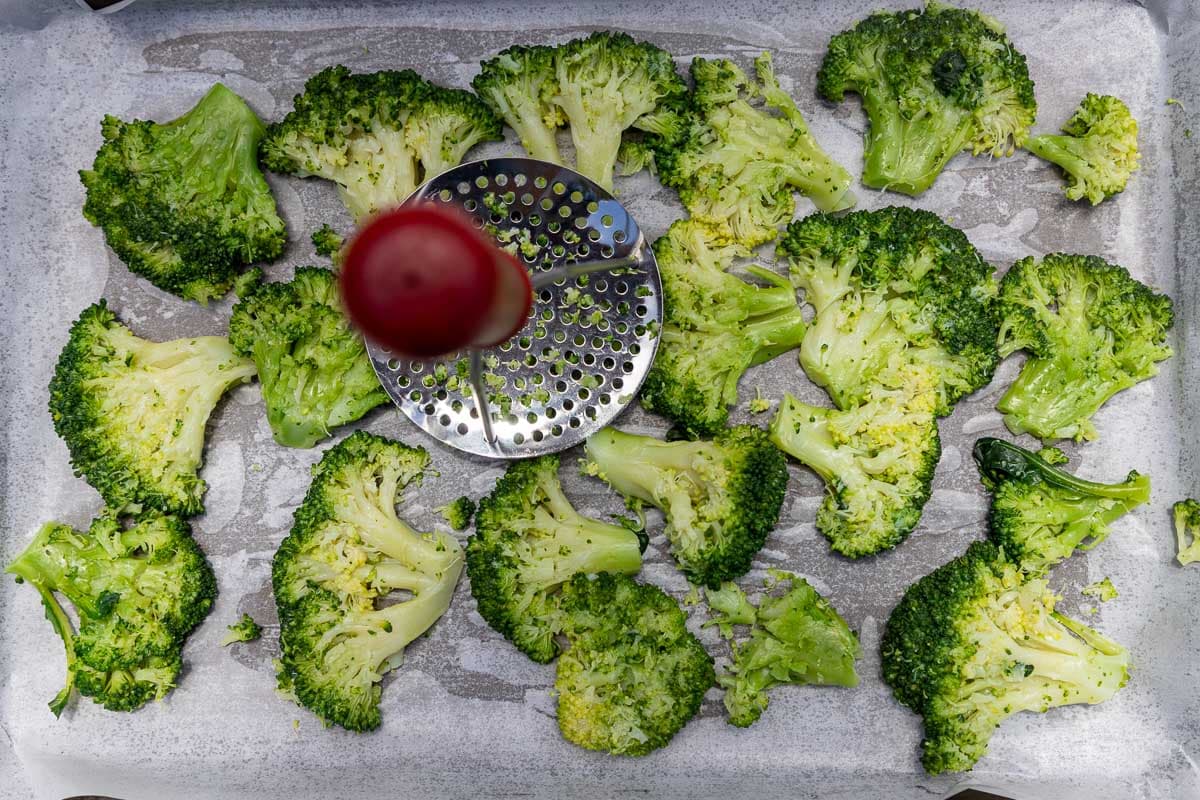 Overhead view of a masher smashing parboiled broccoli on a baking pan lined with baking paper.