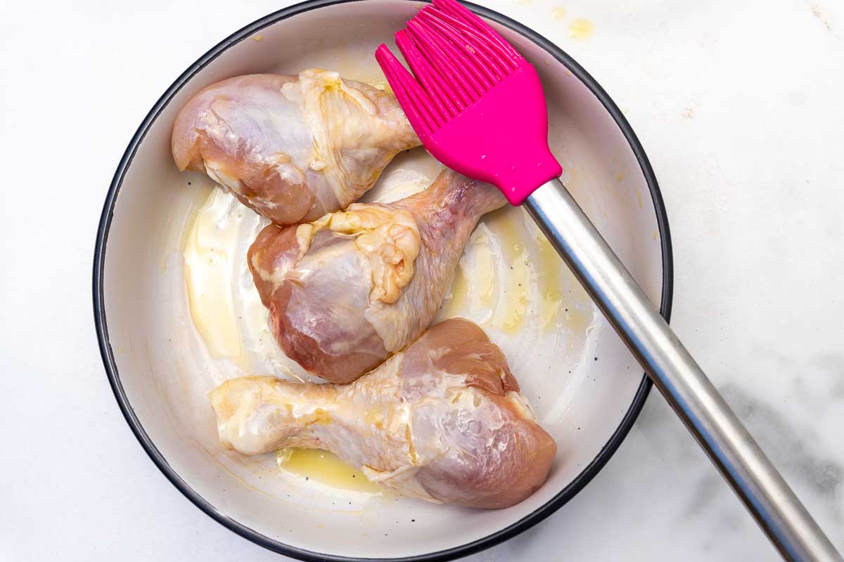 An overhead view of 3 raw chicken legs brushed with olive oil in a white bowl.