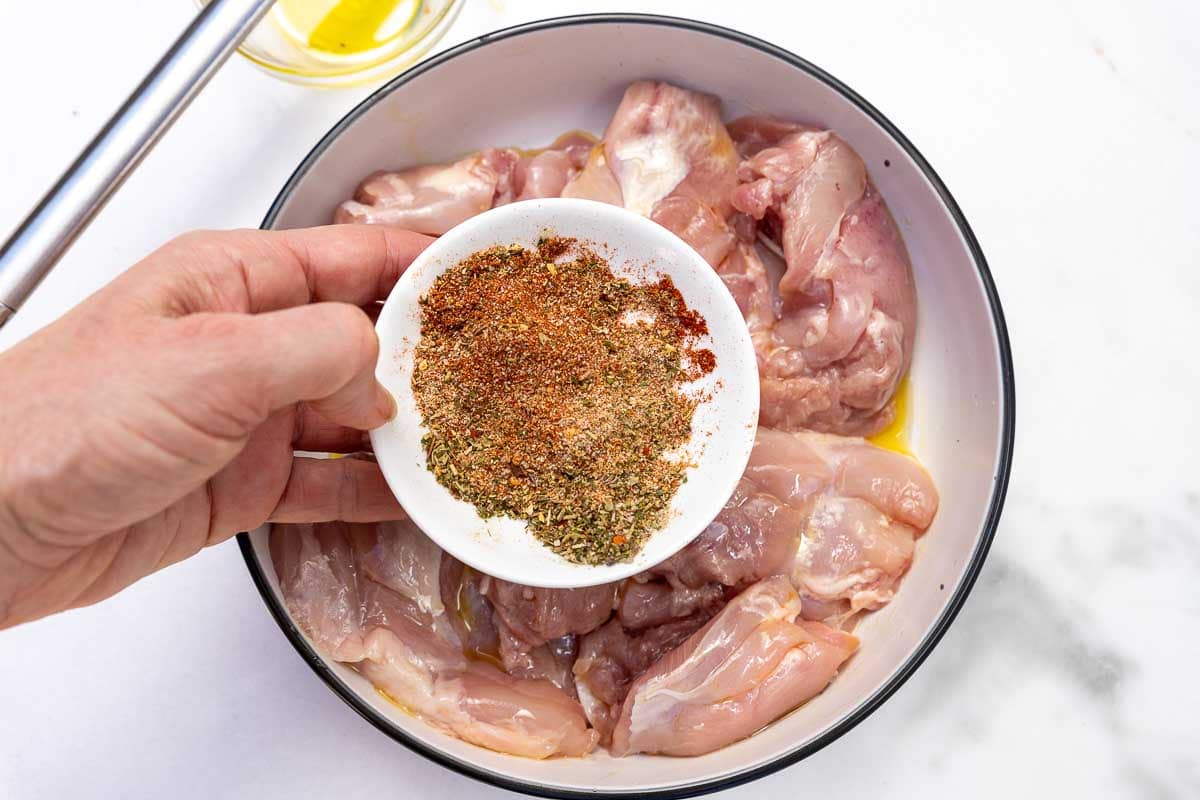 Closeup of someone holding a white bowl of Italian seasoning over a dish of raw chicken thighs brushed with olive oil.