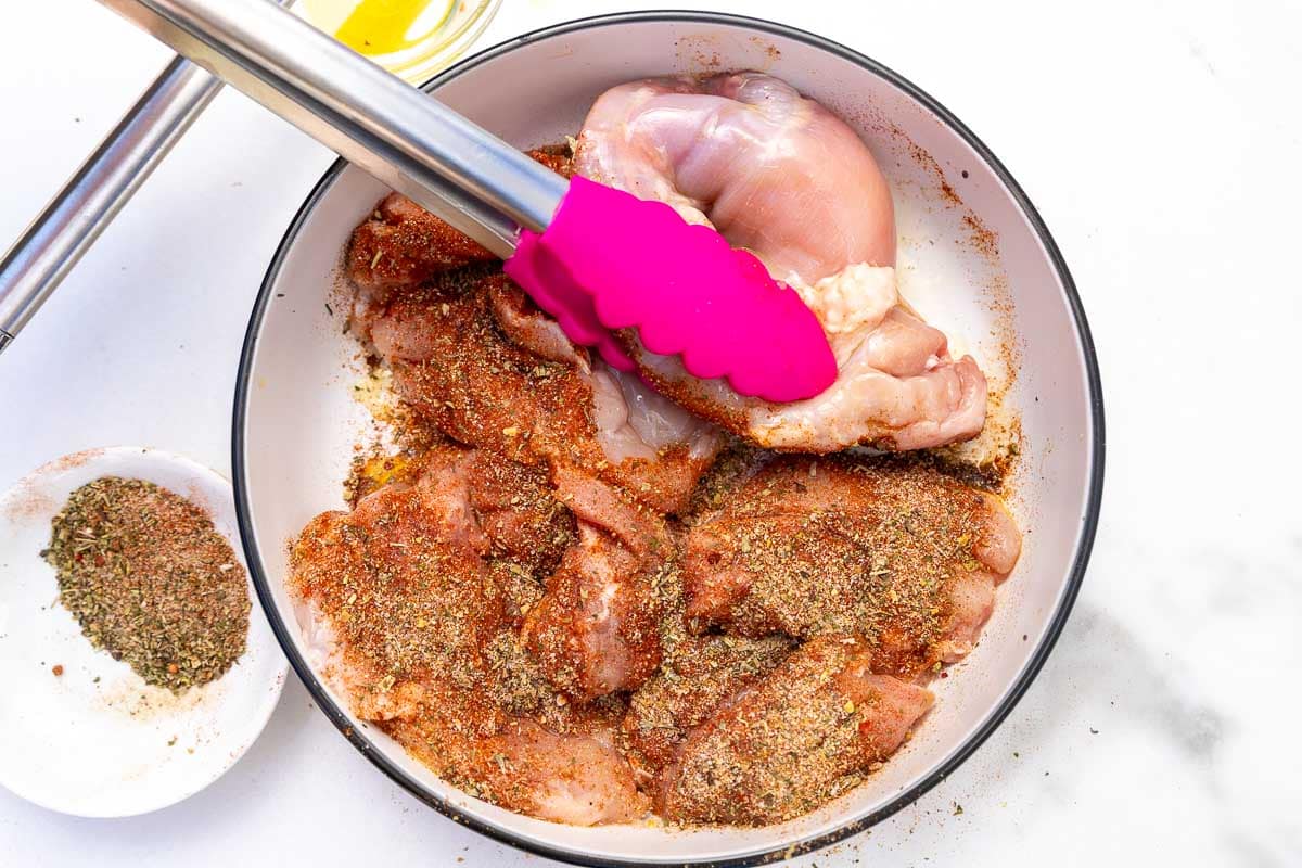 Overhead view of pink tongs turning raw chicken thighs in Italian seasoning in a white bowl.
